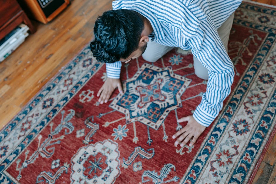 A person kneeling on a patterned red and blue oriental rug in a room with wooden flooring, using a cloth or brush to clean or dust the surface of the carpet. The room features a wooden bookshelf or ca